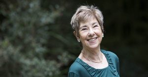Golda Hough stands in front of a dark shrub smiling towars the camera. She is wearing a green top and has short cropped light brown hair. She is smiling brightly.
