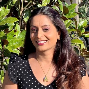 Head and shoulders photograph of Dr Srujana Sahebjada, face to camera, smiling. Dr Sahebjada is standing outdoors. There is a green vine behind her.