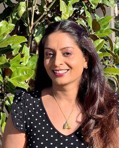 Head and shoulders photograph of Dr Srujana Sahebjada, face to camera, smiling. Dr Sahebjada is standing outdoors. There is a green vine behind her.