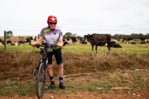Elderly man with a bicycle with a dairy cwo in the background.
