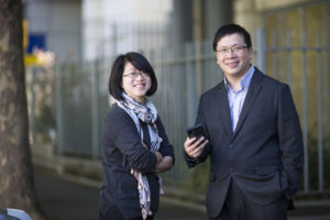 Tena Cheng and George Kong stand together smiling at a camera. George Kong is holding a smart phone.