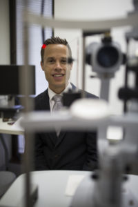Associate Professor Peter van Wijngaarden, sits behind a large clinical eye test machine, as if about to run a test the person behind the camera.