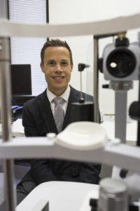 Associate Professor Peter van Wijngaarden, sits behind a large clinical eye test machine, as if about to run a test the person behind the camera.