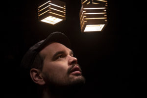 Photo of a man in a cap gazing up at two square wooden lights. The background is dark and his face is illuminated.