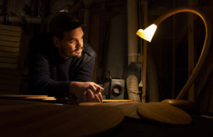 Photo of Duncan Meerding leaning on a desk lift from the side by a tulip shaped lamp on a curved stem. The background is dark.