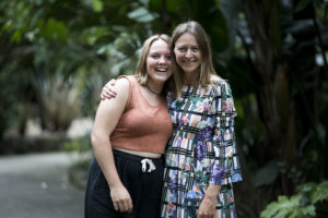 Erica Blake has her arm around her daughter Della. They are smiling and standing in a park surrounded by greenery.