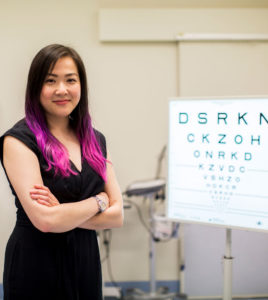 Photograph of Dr Flora Hui standing face to camera in a consultation room with an eye chart to her left hand-side.