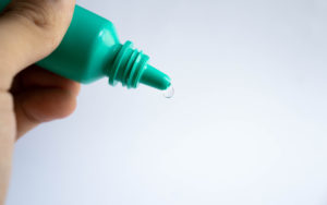 Photograph of a thumb and forefingers holding a green eye drop bottle.