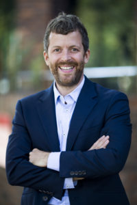 Photograph of Ryan McCarthy, standing outdoors, smiling. He is wearing a blue suit and light blue shirt.