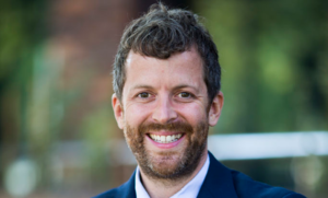 Photograph of Ryan McCarthy, standing outdoors, smiling. He is wearing a blue suit and light blue shirt.