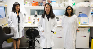 Photograph of Dr Jennifer Fan Gaskin (left), Dr Elsa Chan (centre), and Dr Manisha Shah (right), wearing lab coats, standing and smiling face to camera in a CERA lab, surrounded by equipment.