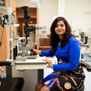 An image of CERA researcher Dr Srujana Sahebjada sitting next to a slit lamp; a common tool used to examine a patient's eye