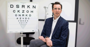 Photograph of Dr Rod O'Day sitting on a chair, face to camera and next to an eye chart in a consultation room.