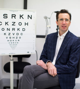 Photograph of Dr Rod O'Day sitting on a chair, face to camera and next to an eye chart in a consultation room.