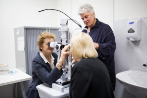A photo of Professor Robyn Guymer and Melina Cain examining a patient. Robyn and the patient are sitting across from one another, while Robyn examines her eyes through a slit lamp. Melinda Cain is standing to the side.