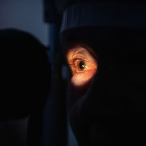 An older women receiving an eye examination. She is shrouded in darkness, except for her eye which is lit up