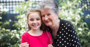 Photograph of clinical research participant Ann and her granddaughter, standing outdoors with greenery in the background. Ann is holding her granddaughter close to her chest, her face against hers. Both are smiling to camera.