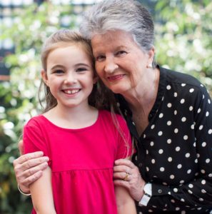 Photograph of clinical research participant Ann and her granddaughter, standing outdoors with greenery in the background. Ann is holding her granddaughter close to her chest, her face against hers. Both are smiling to camera.