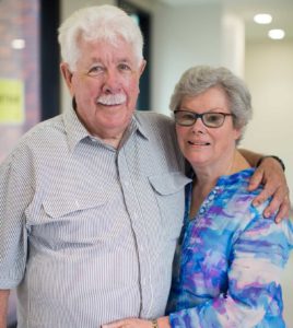Photograph of husband and wife Jim (left) and Clare, standing in a corridor at the Royal Victorian Eye and Ear Hospital, holding one another and smiling face to camera.