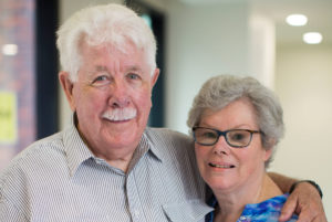 Photograph of husband and wife Jim (left) and Clare, standing in a corridor at the Royal Victorian Eye and Ear Hospital, holding one another and smiling face to camera.