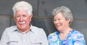 Photograph of husband and wife Jim (left) and Clare, sitting next to each other outside the Royal Victorian Eye and Ear Hospital. Jim is smiling face to camera, and Clare is smiling, looking towards Jim.