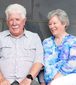 Photograph of husband and wife Jim (left) and Clare, sitting next to each other outside the Royal Victorian Eye and Ear Hospital. Jim is smiling face to camera, and Clare is smiling, looking towards Jim.