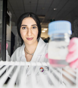 Photograph of CERA PhD Student Layal El Wazan wearing a white lab coat and placing a small specimen jar into a fridge. The camera is placed inside the fridge, pointing outwards, with Layal directly in its field of view, smiling face to camera.