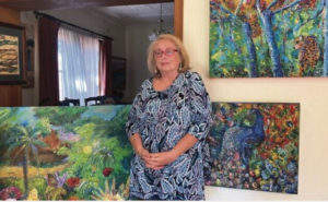 Image of artist Madeleine Popper stanidng in front of three oil paintings. All are brightly colored and have an impressionist style. Madeleine is earing a brightly colored blue patterned dress has shoulder length blond hair and is wearing glasses.