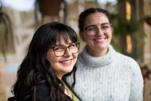 Jasmine Mercieca, smiling, looking over her right shoulder. Her sister Nicole is standing behind her, also smiling, wearing a grey turtleneck jumper.