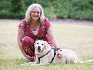 A woman kneeling in the grass with a white Labrador seeing eye dog