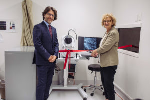Two people standing in front of an ophthalmic imaging machine. One is cutting a bright ribbon across it.