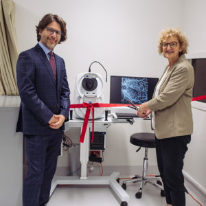 Two people standing in front of an ophthalmic imaging machine. One is cutting a bright ribbon across it.