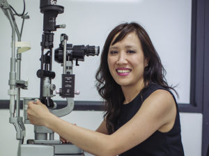 Dr Jennifer Fan Gaskin is sitting at a slit lamp looking towards camera and smiling.
