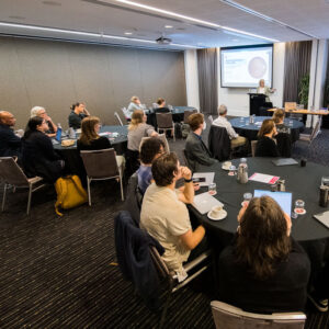 A number of people, sitting around tables in a busy conference room.