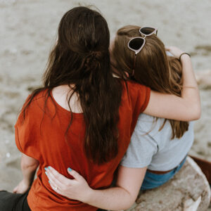 Two young women, sitting on the beach with their arms around one another in support